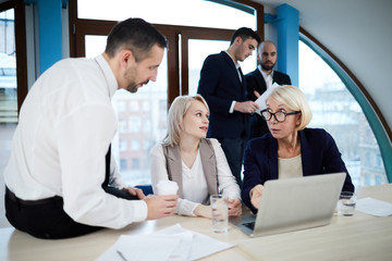 Mature blonde analyst pointing at laptop display and asking question to one of young co-workers at working meeting