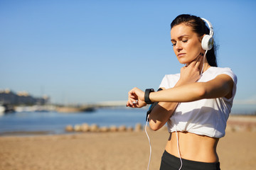 Serious concentrated young female jogger in wired headphones standing on beach and checking pulse...