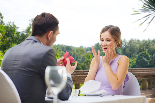 proposal, valentines day and people concept - happy man showing woman present in red box at restaurant over summer background