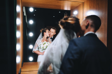 Beautiful newlyweds are standing in the studio opposite the mirror with light bulbs and smiling. Wedding photography. Stylish groom hugging a sweet bride in a lace dress.