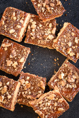 Homemade caramel shortbread squares cookies with nuts laid in the shape of a square on black background with crumbs.