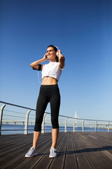 Cheerful confident attractive young female jogger adjusting wired headphones and looking into distance while preparing for training