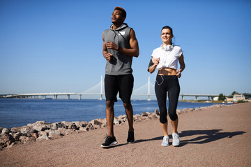Content motivated young multi-ethnic couple in sportswear jogging on city beach while training together in morning