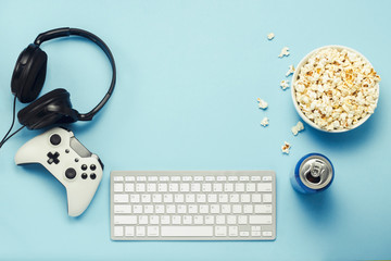 Keyboard and tin can with a drink, energy drink, a bowl of popcorn, a gamepad and headphones on a blue background. The concept of computer games, entertainment, gaming, leisure. Flat lay, top view © Alex