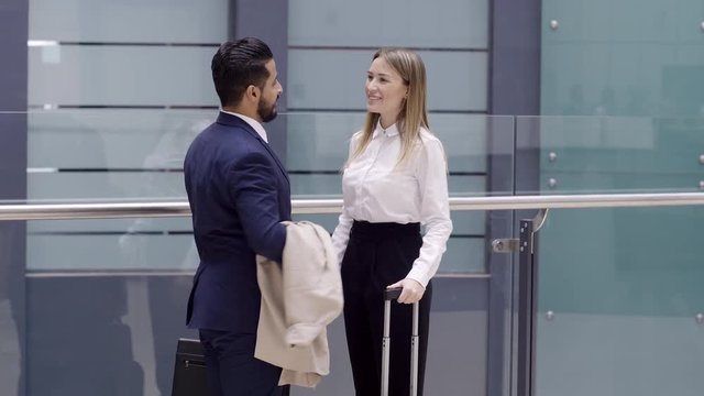 Tilt up shot of Caucasian businesswoman with rolling-bag meeting Arabian businessman in lobby of building, smiling, shaking hands and following him
