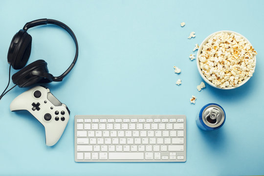 Keyboard and tin can with a drink, energy drink, a bowl of popcorn, a gamepad and headphones on a blue background. The concept of computer games, entertainment, gaming, leisure. Flat lay, top view