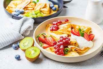 Homemade thin crepes served with curd cream, fruits and berries in black and white plates.