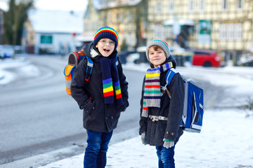 Two little kids boys of elementary class walking to school during snowfall. Happy children having fun and playing with first snow. Siblings ans friends with backpack in colorful winter clothes.