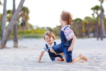 Two little kids boys having fun on tropical beach, happy best friends playing, friendship concept. Siblings brothers, twins fighting, running and jumping in family look with palms trees on background.