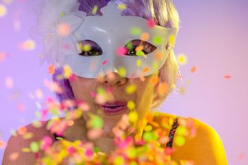 Blowing Confetti. Portrait of latin woman wearing purple wig and makeup mask. Colorful background. Carnival concept, fun and party.