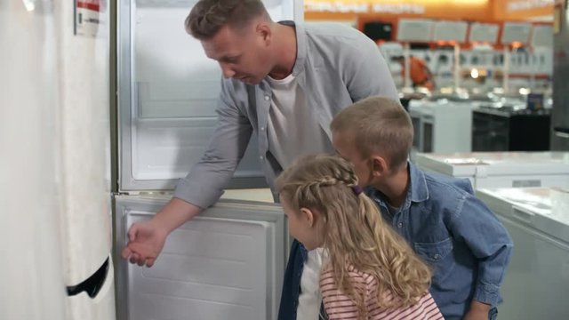 Father, two little daughters and son opening refrigerator and looking inside while shopping together in home appliance store