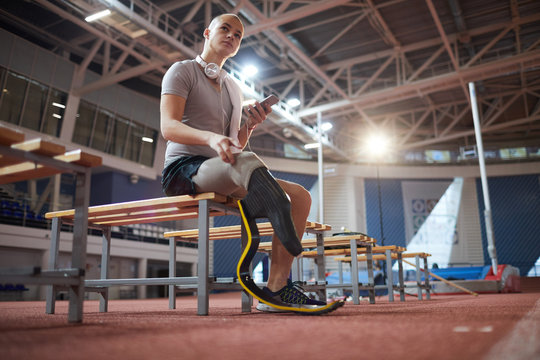 Young pensive sportsman with amputated right leg scrolling in smartphone while resting on bench after training