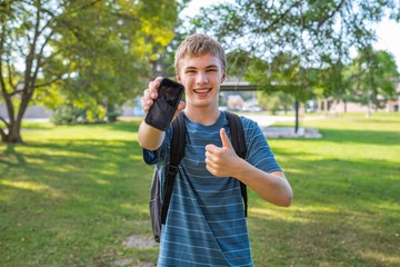 Happy teenager holding his cellphone straight out to display a blank screen. He is standing in a city park on a sunny day.