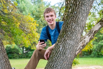 Happy teenager sitting on a tree branch in a park and holding his cell phone.