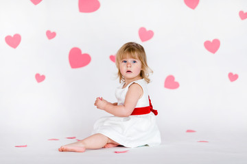 Obraz na płótnie Canvas Little blonde girl in white dress with red ribbon on the white floor with pink hearts on the St. Valentine's day