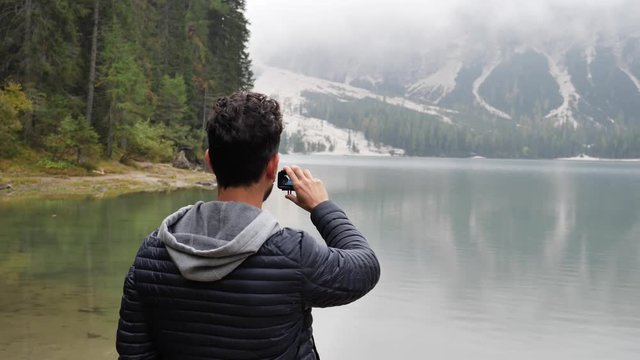 Young handsome man taking photo or recording video with action cam at lake in forest, by the shore. Braies lake or Pragser Wildsee in Trentino region, Italy