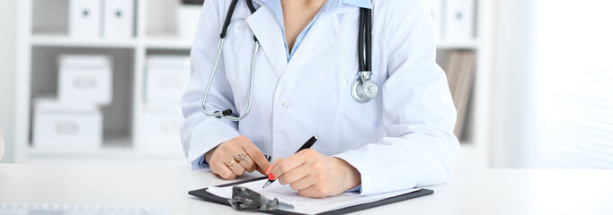 Unknown female doctor working at the desk with clipboard in hospital and filling up medical history...