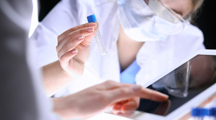 Closeup of scientific research team with clear solution in laboratory. Blonde female chemist holds test tube of glass while her colleague checks results with tablet pc. Blood test, medicine or