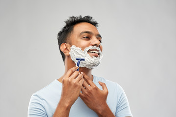 grooming and people concept - young indian man shaving beard with manual razor blade over grey background