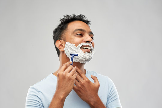 grooming and people concept - young indian man shaving beard with manual razor blade over grey background
