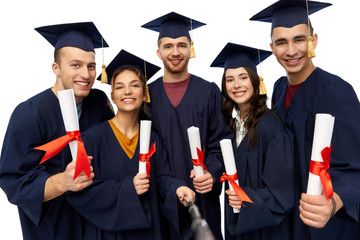 education, graduation and people concept - group of happy graduate students in mortar boards and bachelor gowns with diplomas taking picture by slfie stick over white background