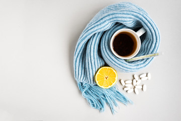 Tea cup with thermometer, blue scarf, pills and lemon on gray background. Flu season, disease.