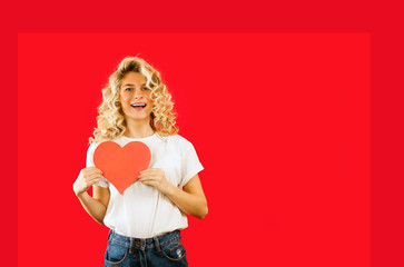 beautiful young emotional girl with a red heart in her hands is standing on an isolated red background.Valentine's Day.