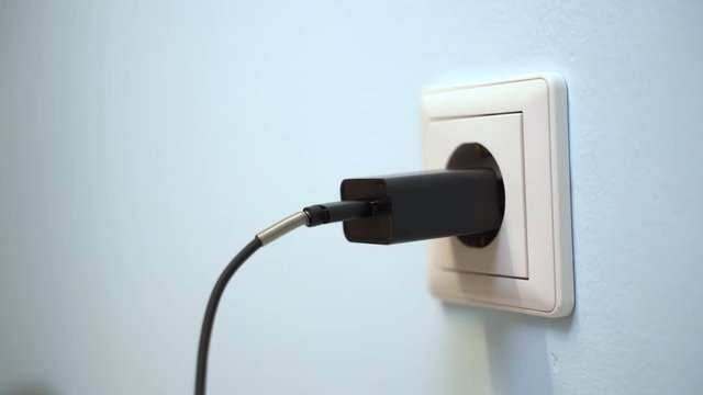 Woman hand plug black phone charger adapter into wall socket.