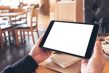 Mockup image of hands holding black tablet pc with blank screen with notebook and bread on wooden table in cafe