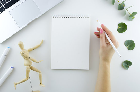 Young woman holds pencil and blank spiral sketchbook on white background. Responsive design template. Space for text Flat lay. Work space with laptop, wooden mannequin, eucalyptus branches and markers