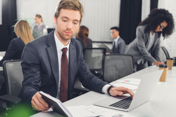 serious young businessman holding notebook and using laptop in office