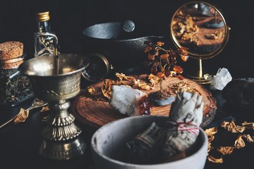 Wiccan witch altar working space. Looks busy and messy filled with dried herbs, citrine crystal, amber decoration, sage stick, gold colored incense burner, gold mirror, mortar and pestle in background