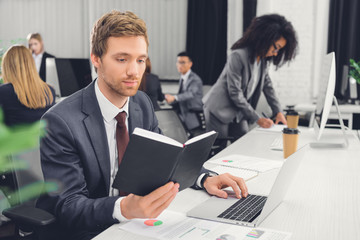 focused young businessman holding notepad and using laptop in open space office