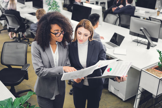 high angle view of young multiracial businesswomen holding papers and discussing project while standing in office