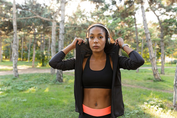 Portrait of sportswoman 20s wearing black tracksuit and headphones, walking through green park