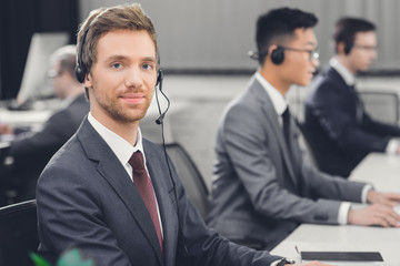 handsome young businessman in headset smiling at camera while working with colleagues in office