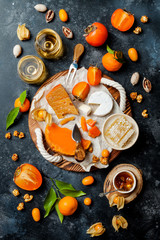 Cheese variety board or platter with cheese assortment, persimmons, honey and nuts. Black stone background. Top view, flat lay