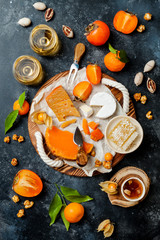 Cheese variety board or platter with cheese assortment, persimmons, honey and nuts. Black stone background. Top view, flat lay