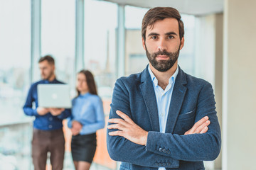 A businessman stands in the office folded his hand to his hand. During this time, he stands against the background of his colleagues who work in the laptop.