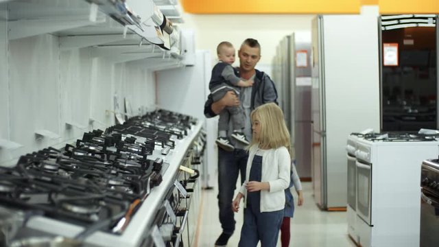 Father holding baby son on arms and walking with two little daughters along home appliance store while choosing kitchen stove