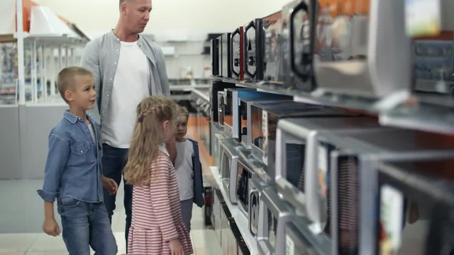 Two little girls and their brother smiling and talking while choosing microwave oven together with dad in home appliance store