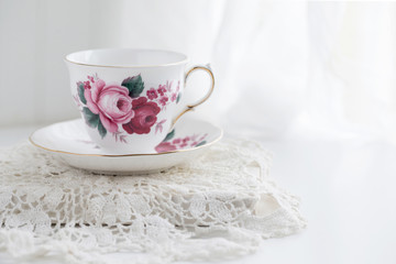 Still life photograph of a white bone china teacup with red roses sitting on a doily covered table against white - obrazy, fototapety, plakaty