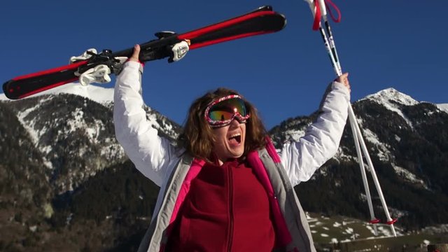 Active girl in a ski suit and mirror glasses with skis and poles in their hands screaming hurray and victory against the background of snowy mountains