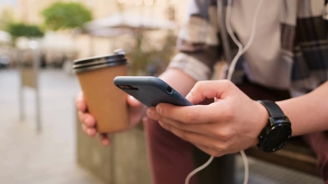 Man using smartphone and drinking coffee to go outdoor. Close-up. Generation z in hipster outfit, using his smartphone, scrolls through social media feed on device, checking map, reading news on app