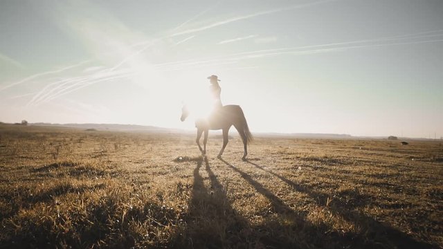 Cowgirl riding brown horse across empty field at sunrise