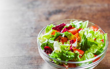 various fresh mix salad leaves with tomato in glass bowl on wooden background.