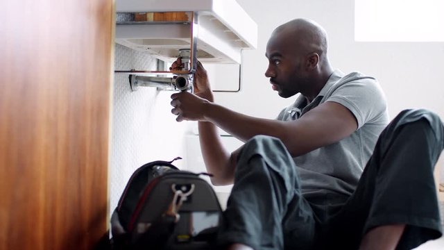 Young black male plumber sitting on the floor and replacing the trap pipe under a bathroom sink, seen from doorway