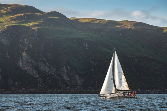 Fototapeta Touristic yacht sailing next to the Northern Ireland steep rocks. Green covered heels, shoreline. Outdoor activity. Perfect background for the creating of various kinds of collages and illustrations.