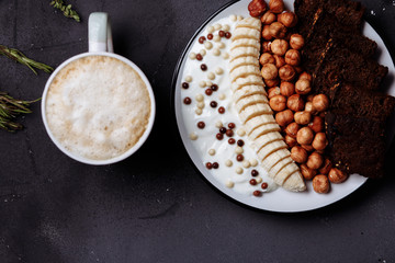 Healthy breakfast with delicious bread, banana, hazelnut, yogurt in bowl and a Cup of coffee, latte or cocoa with milk on a black background, close up