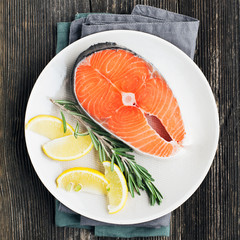 Fresh salmon steak on light plate with lemon and rosemary on wooden background. Top view.